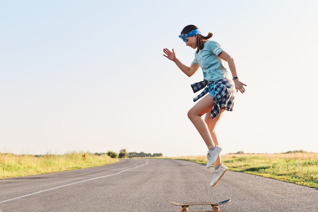 Sporty female wearing t shirt and short doing tricks on a skateboard on street on asphalt road, jumping in the air, enjoying skateboarding alone in sunset in summertime.