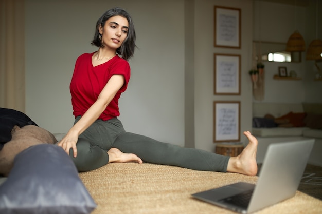 Sporty flexible young woman with canities sitting on floor, strerching legs, doing spinal twist, looking at computer screen, watching online yoga video tutorial with step by step instructions