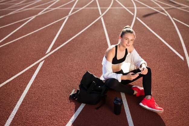 Спортивная девушка в беспроводных наушниках с рюкзаком и спортивной бутылкой рядом мечтательно смотрит в камеру с мобильным телефоном в руках на беговой дорожке стадиона