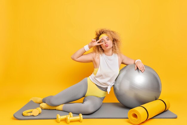 Sporty healthy woman makes peace gesture does sport exercises on fitness mat has workout at home with fitball and dumbbells isolated over yellow background Active lifestyle routine concept