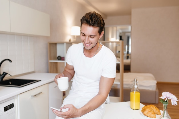 Sporty man smiling while looking at smartphone screen and enjoying tea in morning