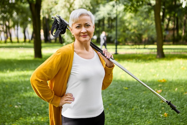 건강한 활동적인 라이프 스타일에 예라고 말하는 은퇴 한 스포티 한 짧은 머리의 여성, 어깨에 노르딕 워킹 스틱을 들고 멋진 산책, 신체 및 심장 혈관계 훈련