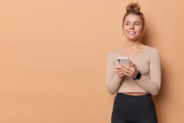 Free photo sporty slim european woman holds mobile phone for messaging and surfing internet dressed in sportswear has satisfied dreamy expression poses against beige background blank copy space for text