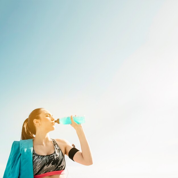 空の背景にボトルから飲むスポーティな女性