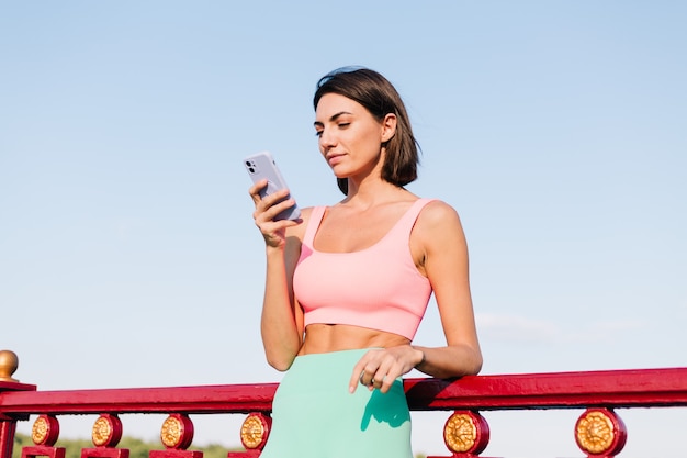 Спортивная женщина в подходящей спортивной одежде на закате на современном мосту с видом на реку счастливая позитивная улыбка с мобильным телефоном смотрит на экран