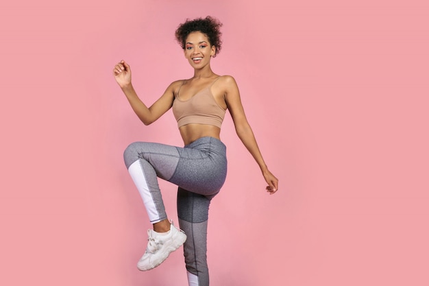 Foto gratuita la donna sportiva che pratica lo squat si esercita in studio. donna africana in abiti sportivi che risolve su sfondo rosa.