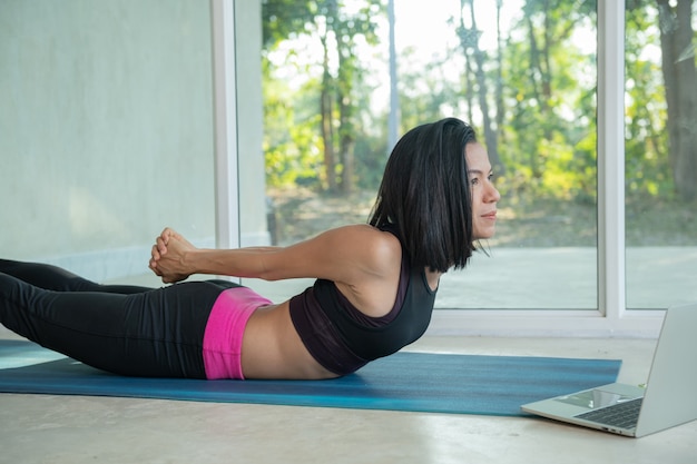 Спортивная женщина практикует йогу, тренирует позу спины и ягодиц, растягивает, смотрит видеоурок по фитнесу онлайн на ноутбуке, делает тренировку дома, сидя на коврике в гостиной, практикуя