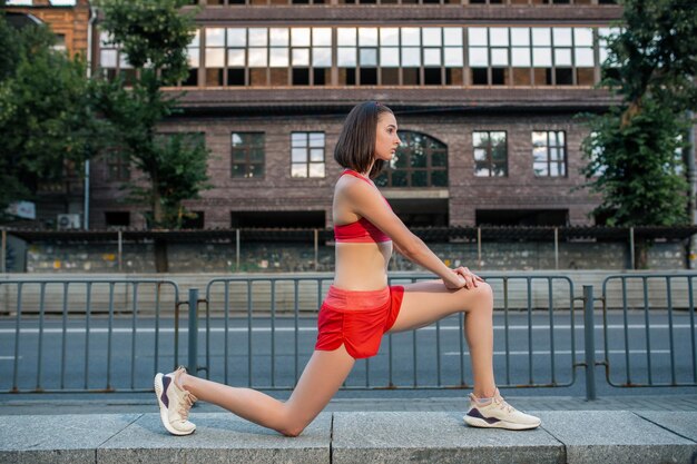 都会のフィットネストレーニングを実行する前に、スポーティな女性が足を伸ばしてウォーミングアップします。スポーツと健康的なライフスタイルのコンセプト。外で運動する女性アスリート。