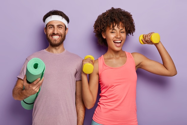 Sporty woman trains with dumbbells, has cheerful look, her husband stands near, holds rolled up fitness mat, isolated on purple background