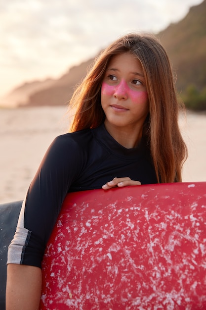 Sporty woman with surf zinc, dressed in black wetsuit, holds waxed surfboard