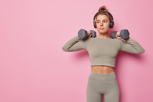 La giovane donna sportiva fa esercizi con manubri bodybuilding allenamento essendo in buona forma fisica vestita con top corto e leggings ascolta musica tramite cuffie isolate su sfondo rosa