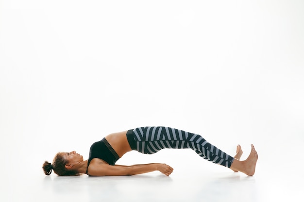 Sporty young woman doing yoga practice isolated on white wall. Fit flexible female model practicing. Concept of healthy lifestyle and natural balance between body and mental development.