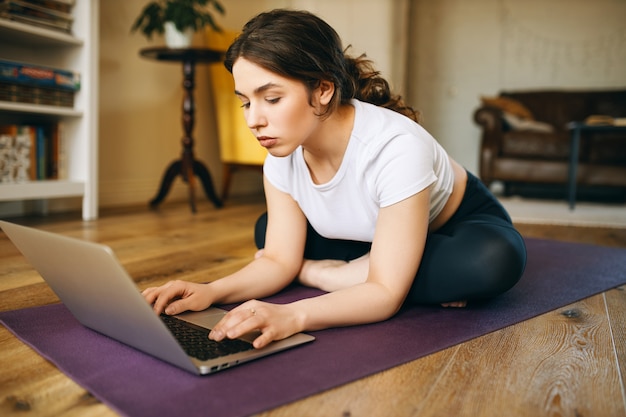 실내 매트에서 운동하는 동안 먼 거리에서 작동하는 열린 노트북 keyboarding 앞에 크로스 legged 앉아 레깅스에 스포티 한 젊은 여자. 사람, 기술, 커뮤니케이션 및 피트니스 개념