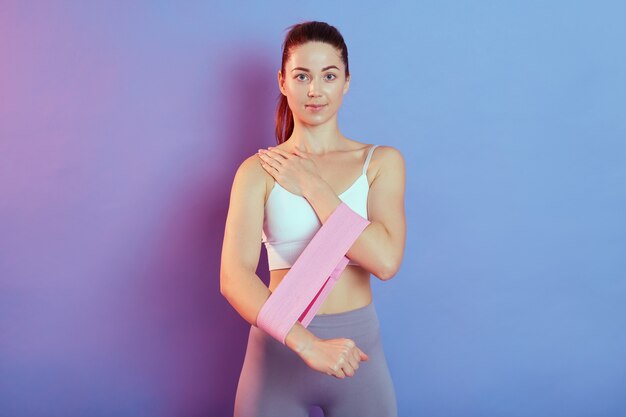 Sporty young woman model wearing white sleeveless t shirt and leggins, doing sport exercise on biceps and triceps with sport fitness rubber bands isolated on color background.