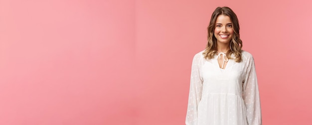 Spring beauty and women concept Portrait of tender feminine blonde girl with white beaming smile standing in light cute dress over pink background with happy attitude positive emotions