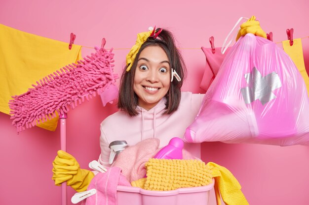 Spring cleaning concept. Positive Asian housewife holds mop bag of cleaning detergents does laundry at home cleans room poses against clothes hanging on clothesline with clothespins. Cleanup concept