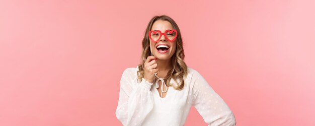 Spring happiness and celebration concept Closeup portrait of funny and carefree beautiful caucasian woman with blond hair white dress holding heartshaped glasses mask and laughing