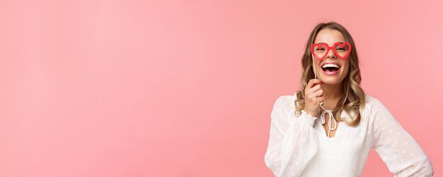 Spring happiness and celebration concept Closeup portrait of funny and carefree beautiful caucasian woman with blond hair white dress holding heartshaped glasses mask and laughing