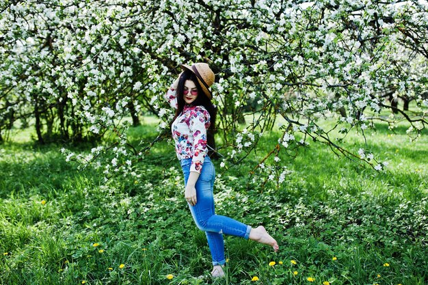 Spring portrait of brunette girl in pink glasses and hat at green blossom garden