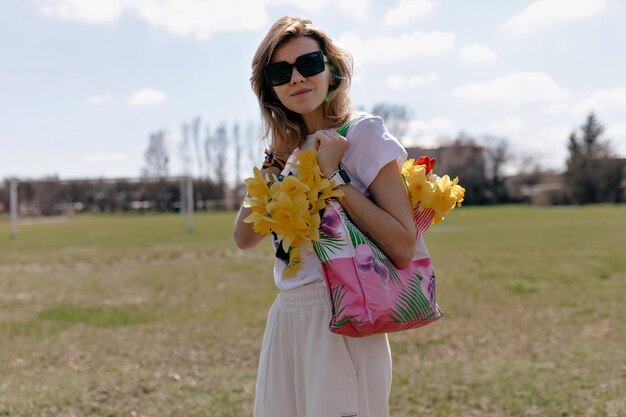 Spring portrait of charming young girl on the green field Lovely woman with short light hairstyle is holding bright bag with flowers touching head and smiling
