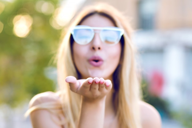 Sprint positive portrait of happy cheerful young woman sending air kiss to you , wearing clear glasses, focus on hand, pastel toned colors.