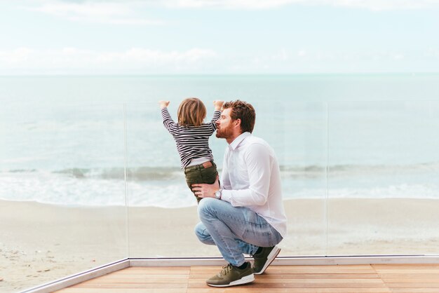 Squatting man holding curious toddler on seashore