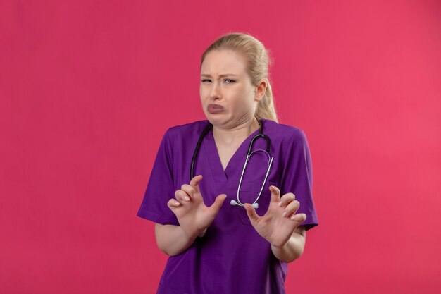 Squeamish doctor young girl wearing purple medical gown and stethoscope on isolated pink background