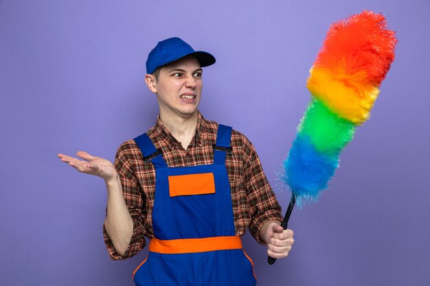 Squeamish young cleaning guy wearing uniform and cap holding and looking at pipidastre 