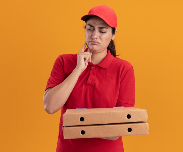 Ragazza di consegna giovane schizzinosa che indossa l'uniforme e la tenuta del cappuccio e guardando le scatole per pizza mettendo il dito sulla guancia isolata sulla parete arancione