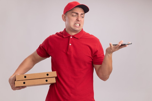 Squeamish young delivery man wearing uniform with cap holding pizza boxes and looking at phone in his hand isolated on white wall