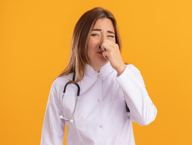 Squeamish young female doctor wearing medical robe with stethoscope grabbed nose isolated on yellow wall