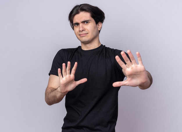 Squeamish young handsome guy wearing black t-shirt showing stop gesture isolated on white wall