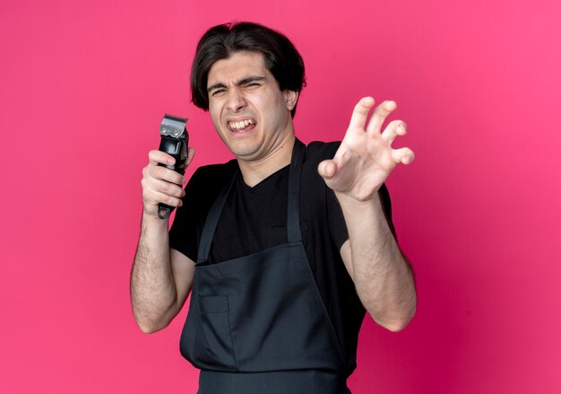 Squeamish young handsome male barber in uniform holding hair clippers and showing tiger style gesture isolated on pink