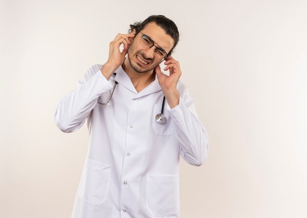 Squeamish young male doctor with optical glasses wearing white robe with stethoscope closed ears on isolated white wall with copy space