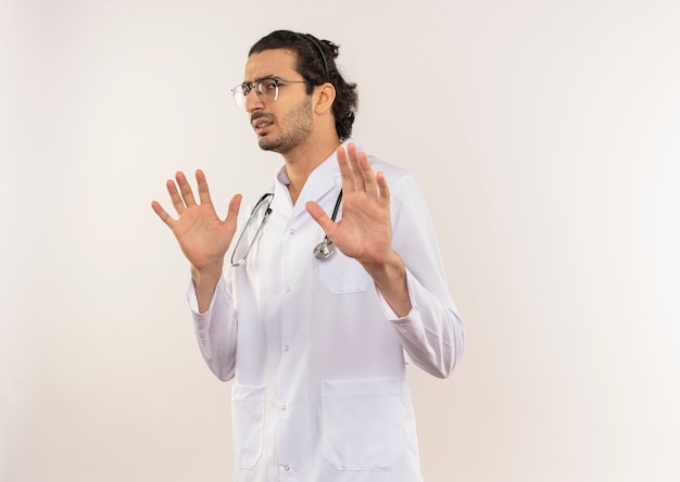 Squeamish young male doctor with optical glasses wearing white robe with stethoscope raising hands on isolated white wall with copy space