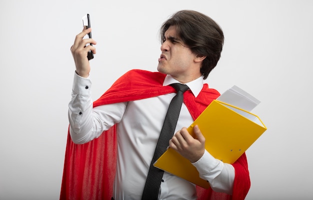 Бесплатное фото Брезгливый молодой супергерой в галстуке держит папку и смотрит на телефон в руке, изолированной на белом фоне