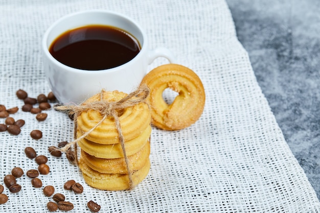 Pila di biscotti con chicchi di caffè e una tazza di caffè su una tovaglia bianca.