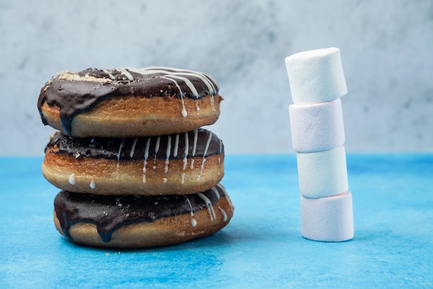 Stack of chocolate donuts and marshmallows on blue table. 