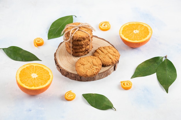 Stack of homemade cookie on wooden board and half cut orange with leaves over white surface.