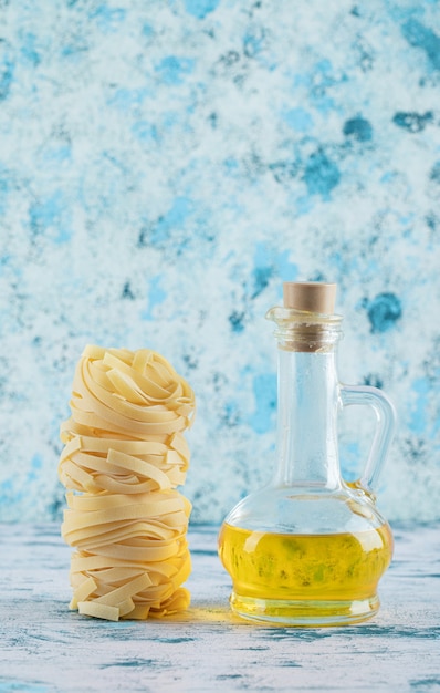 Stack of tagliatelle nests and glass of olive oil on blue.