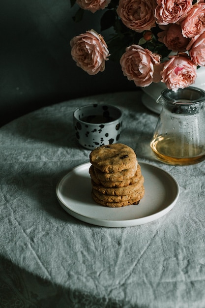 Stacked baked cookies on a plate near a cup and teapot, and pink roses in a vase on a table