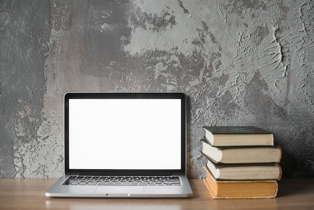 Stacked books and laptop with blank white screen on wooden surface