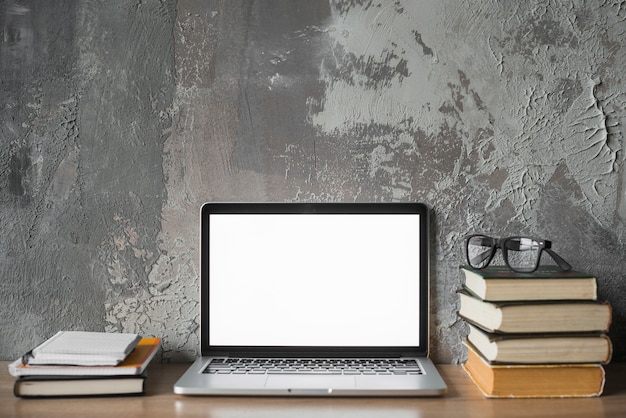 Stacked books; spectacles and laptop with blank white screen on wooden surface