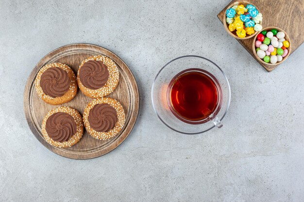 Сложенное печенье и две миски с конфетами на деревянных досках вокруг чашки чая на мраморной поверхности.