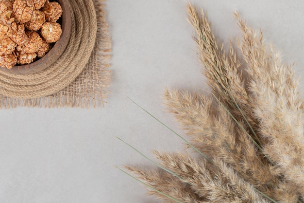 Stalks of dried needlegrass next to a wooden bowl on trivet, filled with candied popcorn on marble background. 