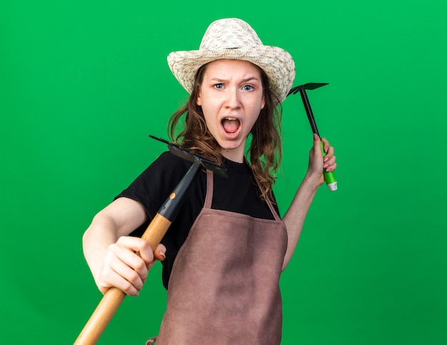 Стоя в боевой позе, молодая женщина-садовник в садовой шляпе держит грабли с мотыгой
