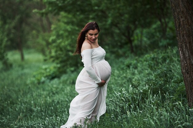 Standing in the grass. Beautiful pregnant woman in dress have a walk outdoors. Positive brunette