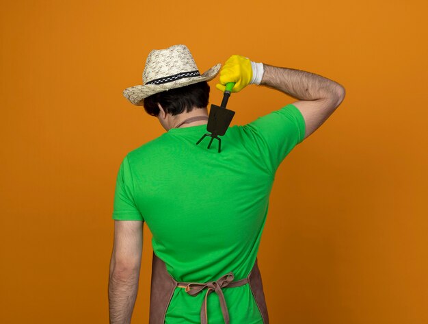 無料写真 オレンジ色に分離されたくわ熊手で肩を引っ掻く手袋とガーデニング帽子を身に着けている制服を着た若い男性の庭師の後ろに立って