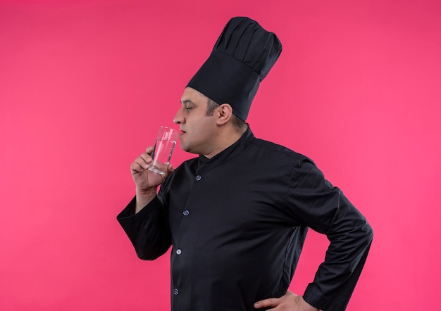 Standing in profile view middle-aged male cook in chef uniform drinking water from glass with copy space