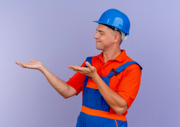 Стоя в профиль, довольный молодой мужчина-строитель в униформе и защитном шлеме, притворяющийся, что держит что-то на фиолетовом